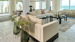 A spacious living room with a large sofa, a coffee table, and a chandelier. There is a view of the ocean through large windows.