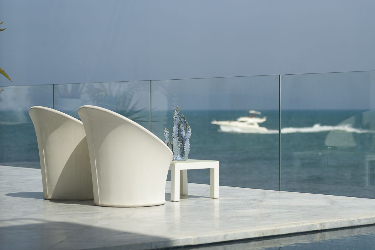 Un área de estar moderna al aire libre con dos sillas blancas y una mesita, con vistas a un mar tranquilo donde se divisa un barco a lo lejos.