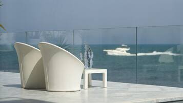 Un área de estar moderna al aire libre con dos sillas blancas y una mesita, con vistas a un mar tranquilo donde se divisa un barco a lo lejos.