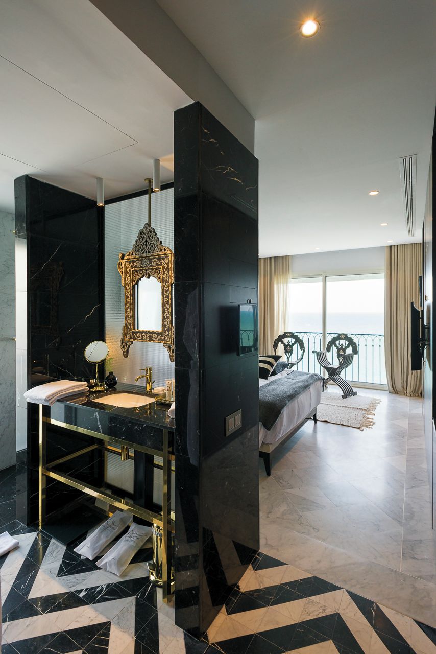 A modern hotel room with a large bed, a bathroom with a double sink, and a large window offering a view.
