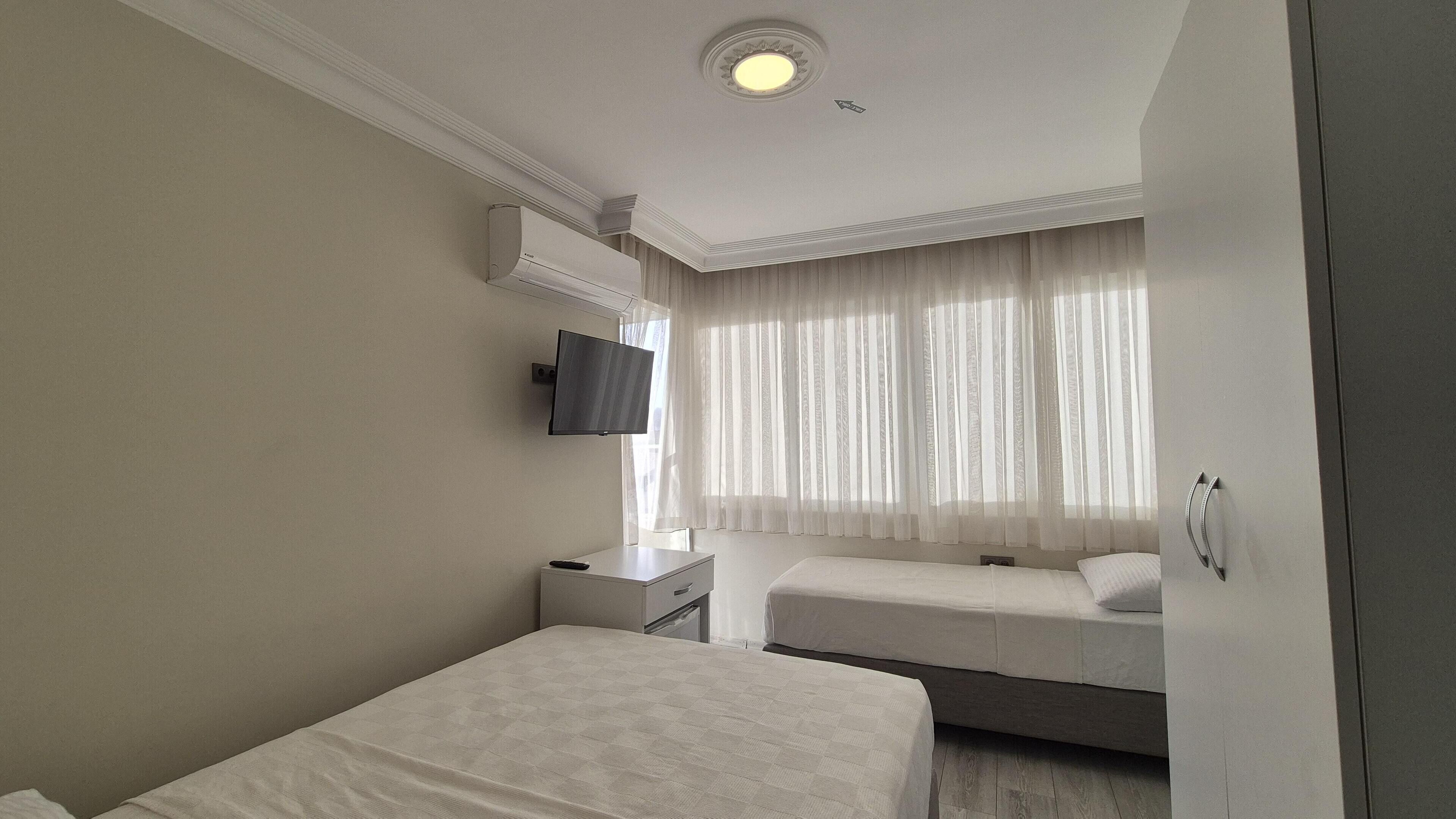 Habitación de hotel con cama, mesita de noche, televisor montado en la pared y ventana con cortinas translúcidas.