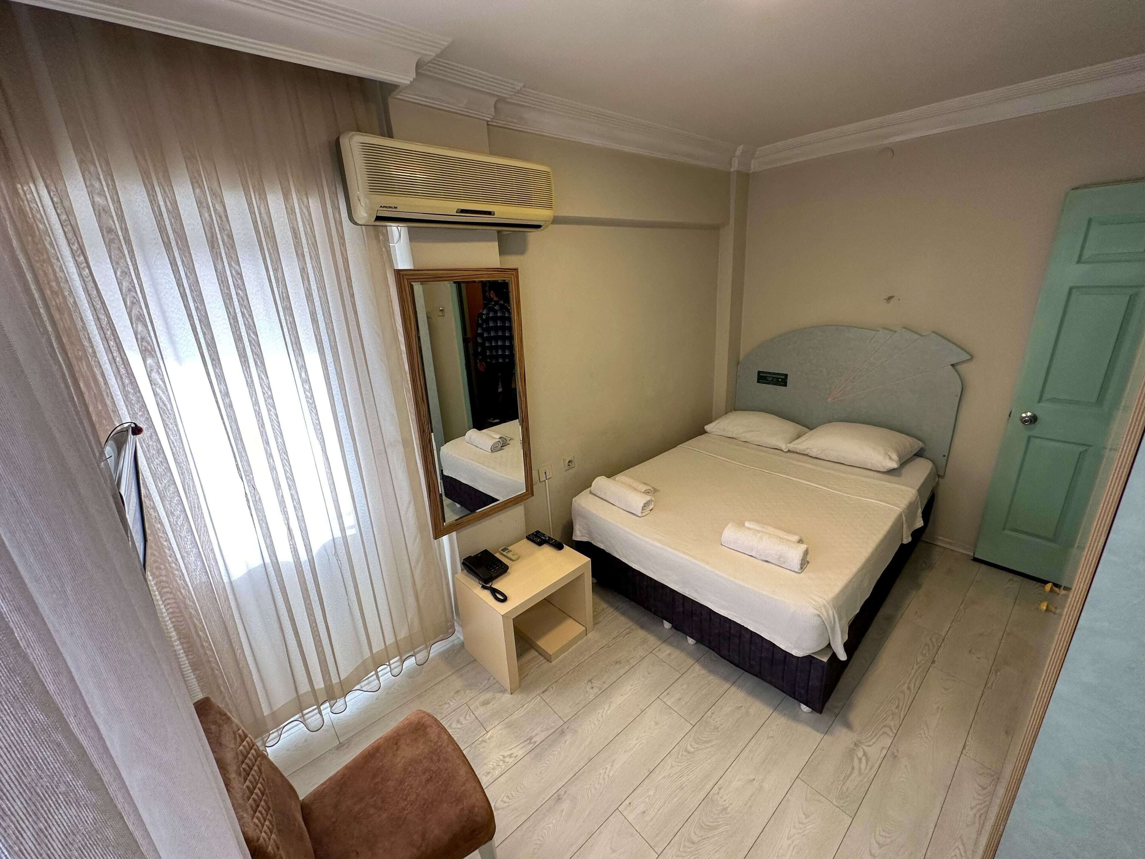 A hotel room with a bed, a bedside table, a mirror, an air conditioning unit, and a door.