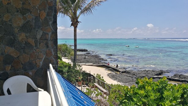A stone wall, a white plastic chair, a blue awning, and a view of the ocean with boats. - Blue Beryl Guest House (Blue Bay)