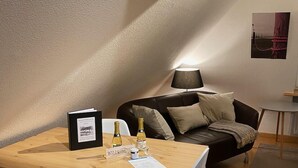 Une pièce chaleureuse avec une table en bois, un canapé en cuir noir et une petite table blanche. On y trouve deux bouteilles, un tableau encadré et une plaque nominative.