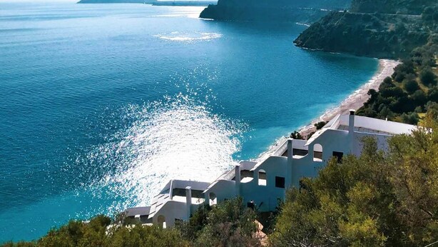 A coastal view with a beach, clear blue sea, and white buildings with balconies overlooking the water.