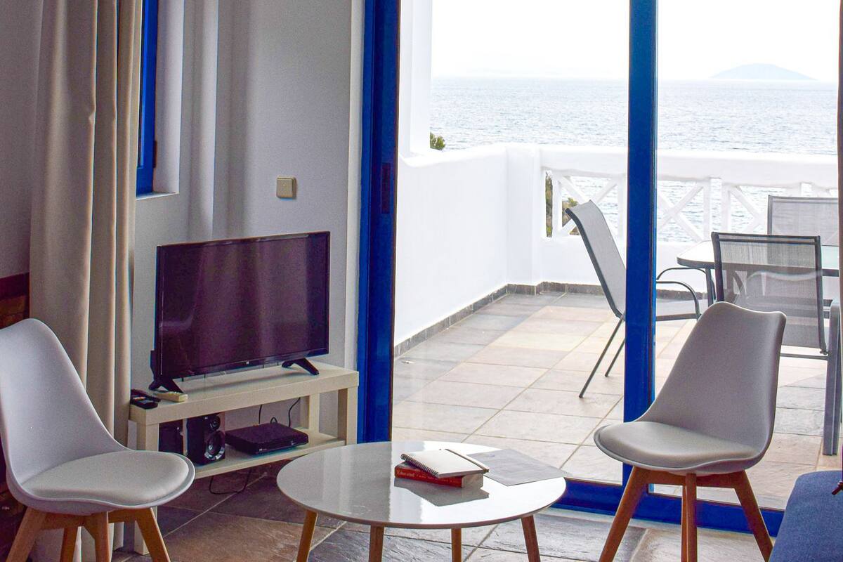 Ett rum med en TV, två stolar, ett litet bord och utsikt över havet genom en dörr med blå ram.