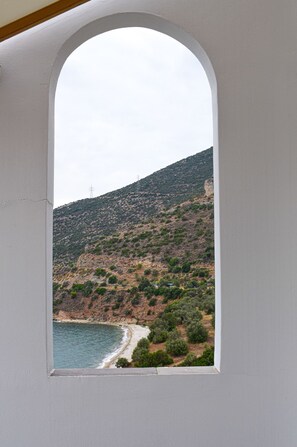 A coastal landscape through a window with a white frame, featuring a beach, rocky hillside, and sparse vegetation. - Muses Seaside Villas (North Kynouria)