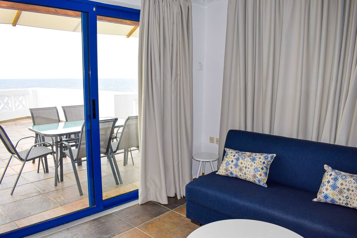 Ett rum med en blå soffa, ett glasbord med stolar och utsikt över havet genom en glasdörr.