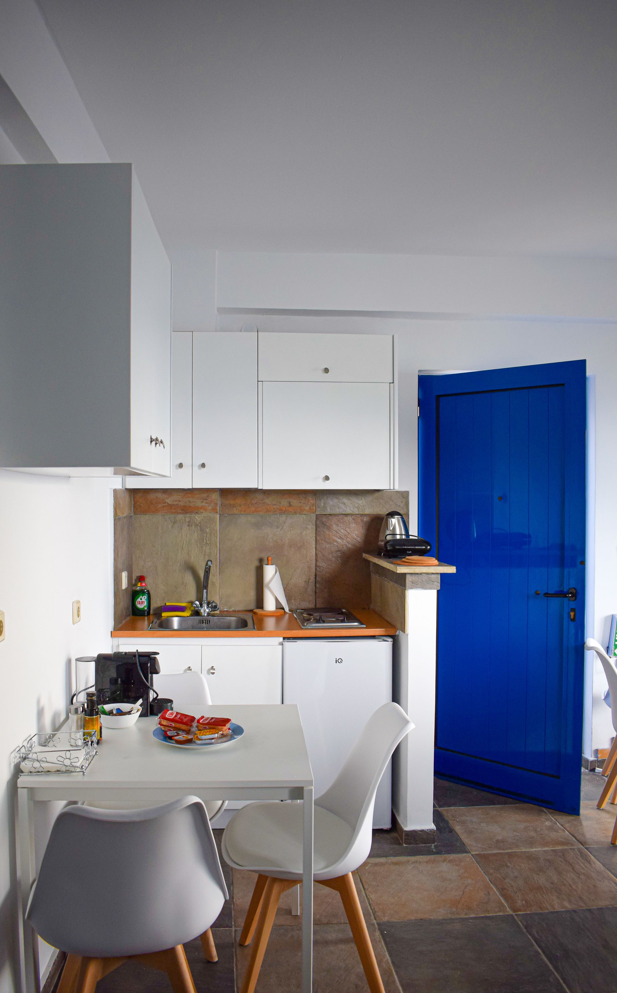 Una cocina compacta con armarios blancos, una puerta azul y un comedor con una mesa y sillas blancas.