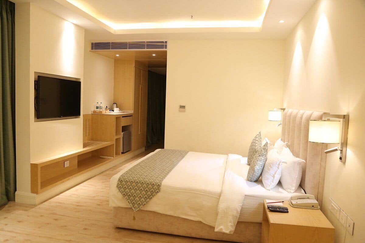 Habitación de hotel con cama, mesita de noche, lámpara y televisor montado en la pared.