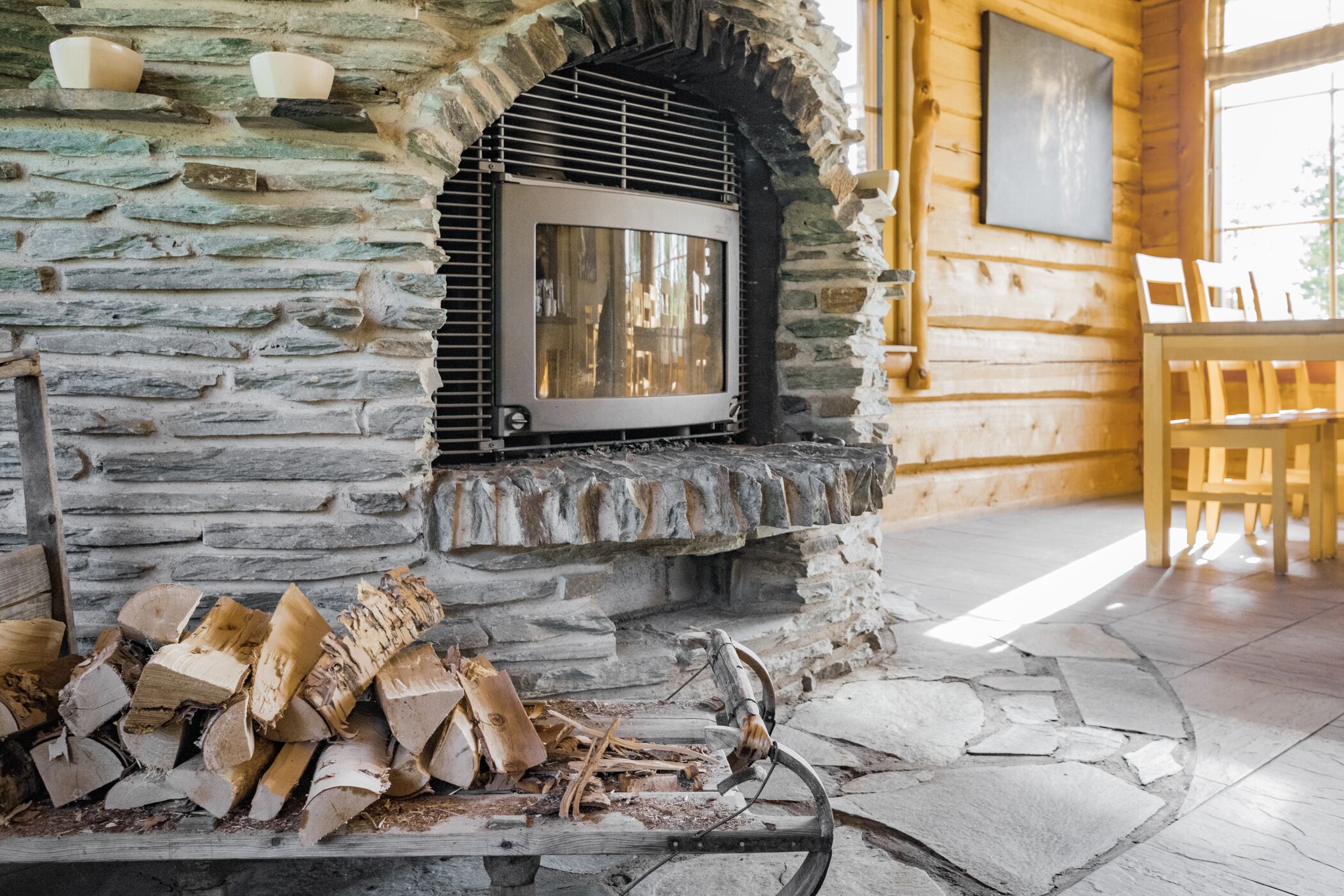 A stone fireplace with a TV, wood logs, and a dining area with wooden chairs.