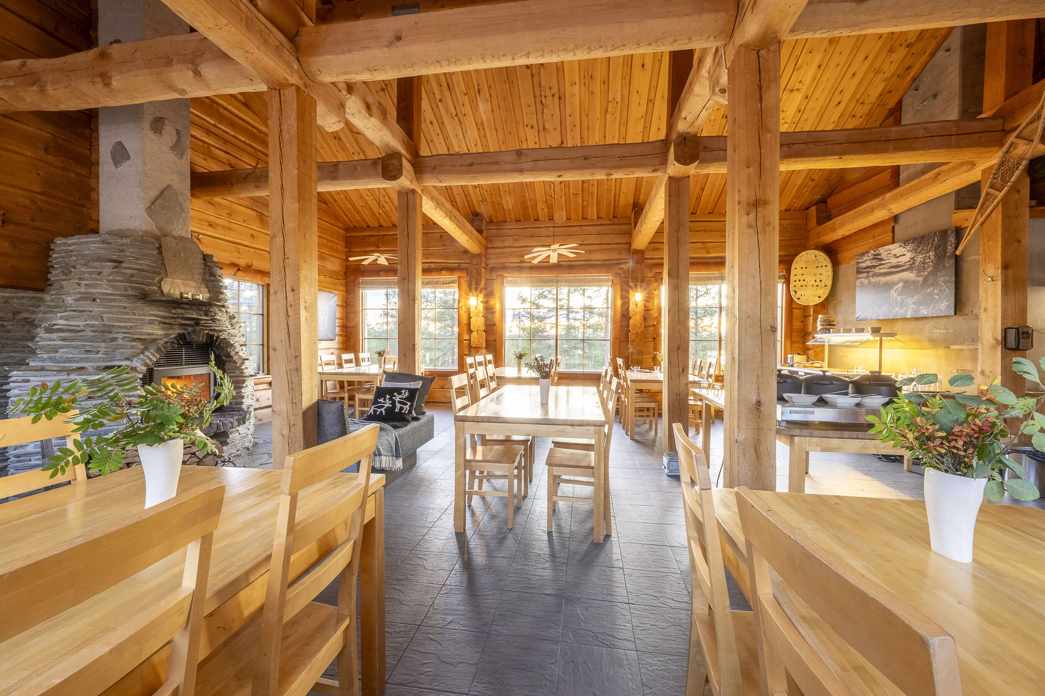 A rustic wooden interior with a stone fireplace, wooden tables and benches, and large windows allowing natural light.