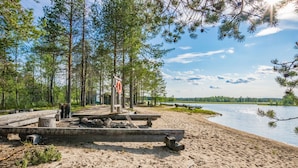 Hiekkainen ranta, jossa on puinen laituri, ja sen ympärillä kohoaa korkeita puita sekä levittäytyy tyyni järvi.