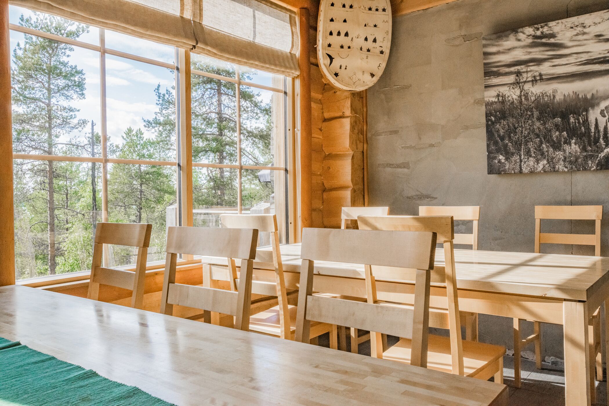 A wooden dining table with chairs, large windows, and a painting on the wall.
