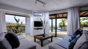 A spacious living room with a large TV, comfortable seating, and a view of the pool and desert landscape. - Villa Thea (Pegeia)