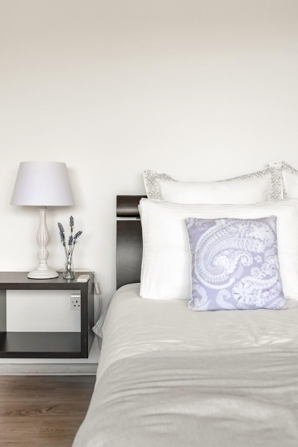 A neatly made bed with white bedding and a patterned pillow, a bedside table with a lamp and a vase of flowers, and a power outlet on the nightstand.