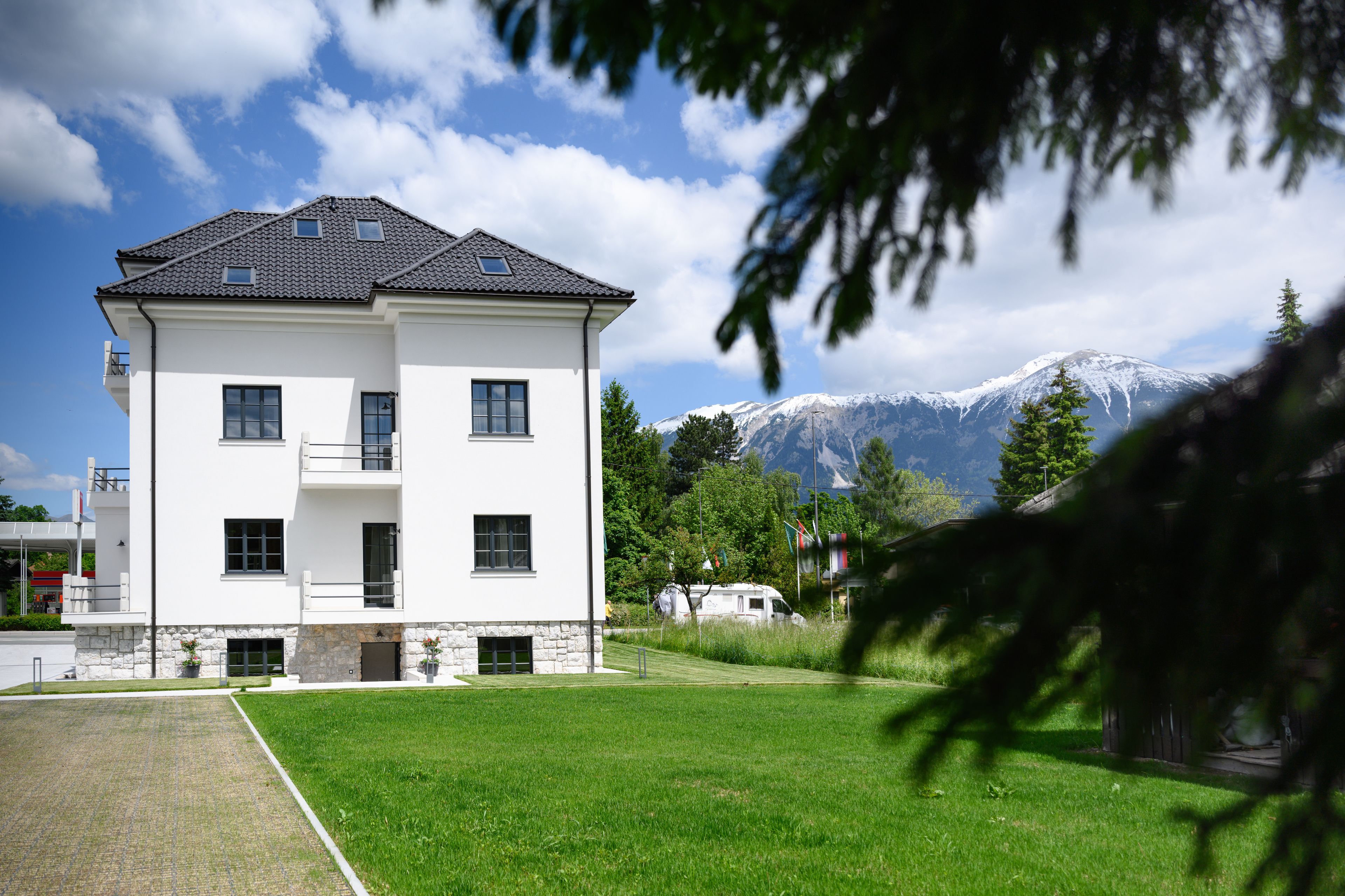 A two-story white building with a stone base, a balcony, and a view of snow-capped mountains.