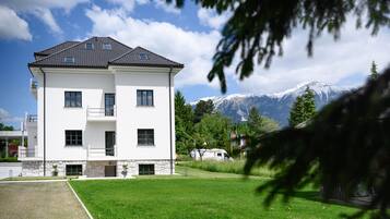A two-story white building with a stone base, a balcony, and a view of snow-capped mountains.