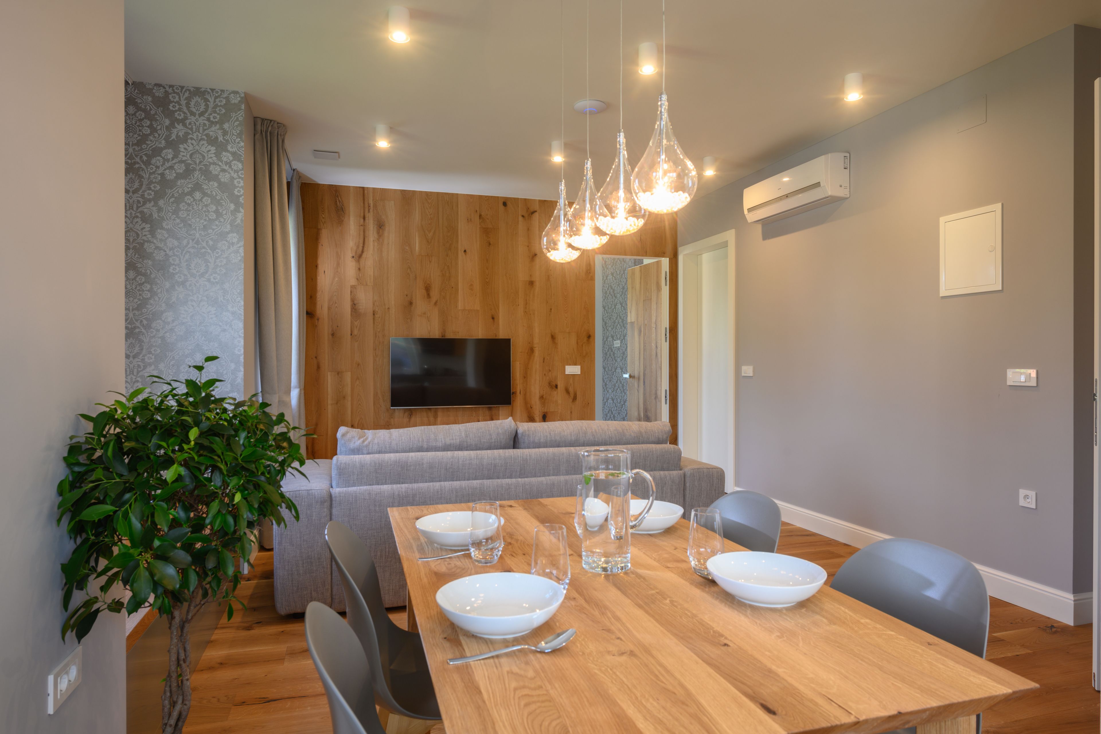 Un espace repas moderne avec une table en bois dressée pour quatre personnes, un canapé gris et un téléviseur fixé au mur.