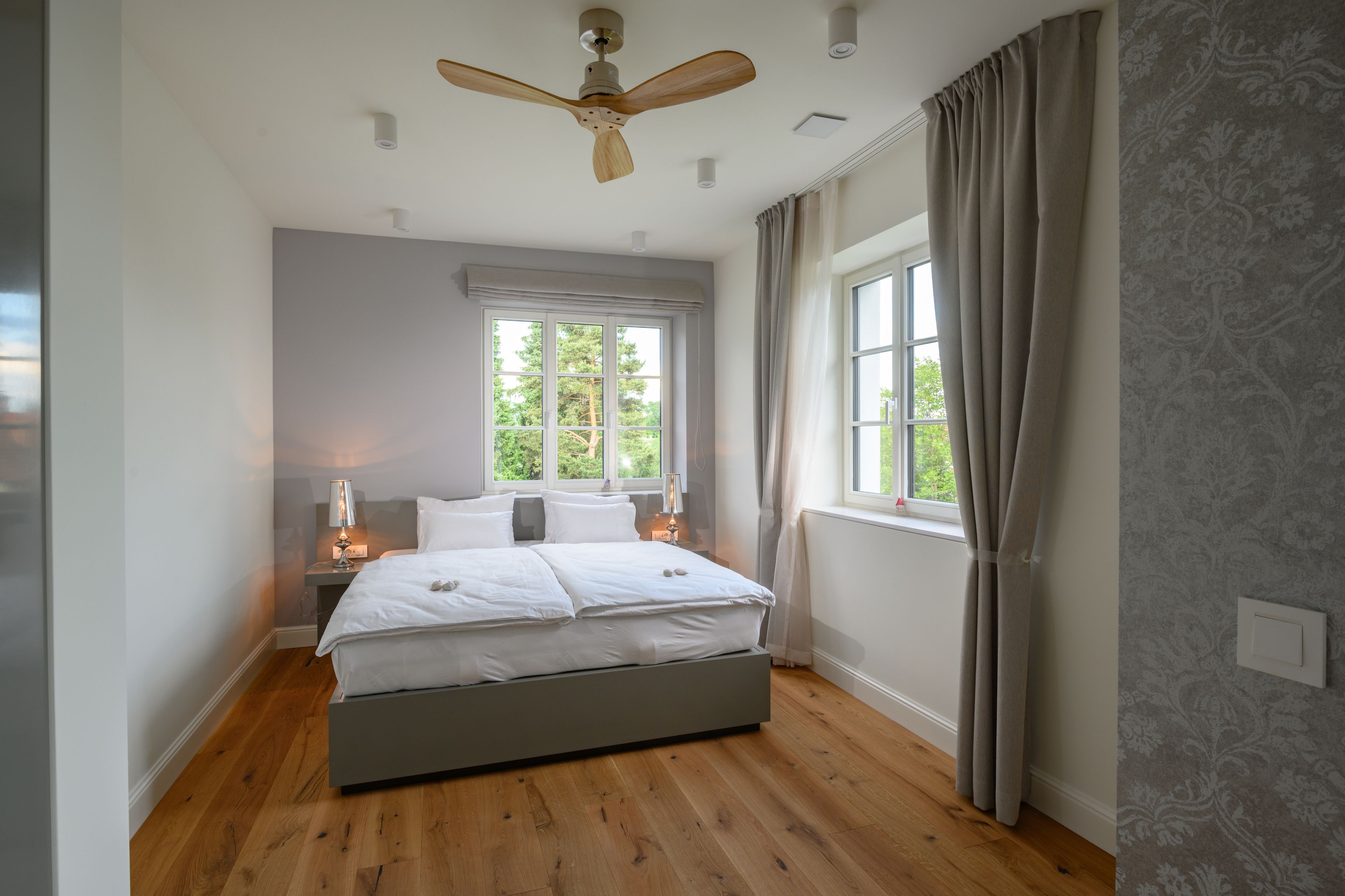 Un dormitorio con cama, ventilador de techo, ventana con cortinas y mesita de noche con lámpara.