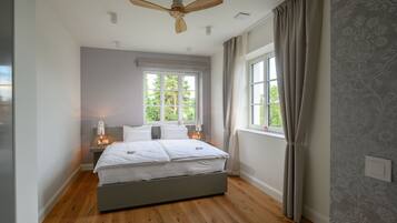 Un dormitorio con cama, ventilador de techo, ventana con cortinas y mesita de noche con lámpara.