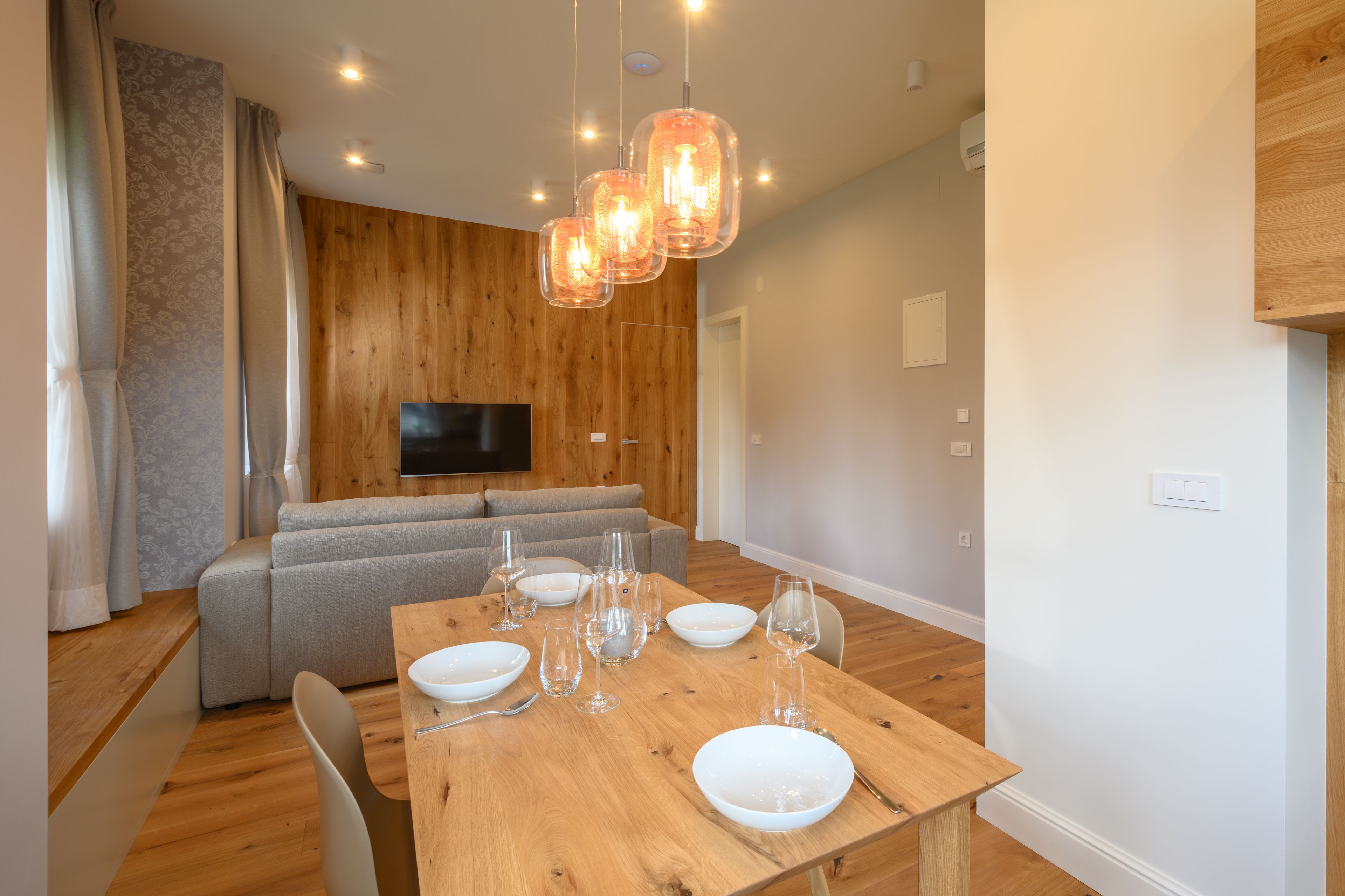A dining area with a wooden table set for four, a grey sofa, and a TV mounted on the wall.