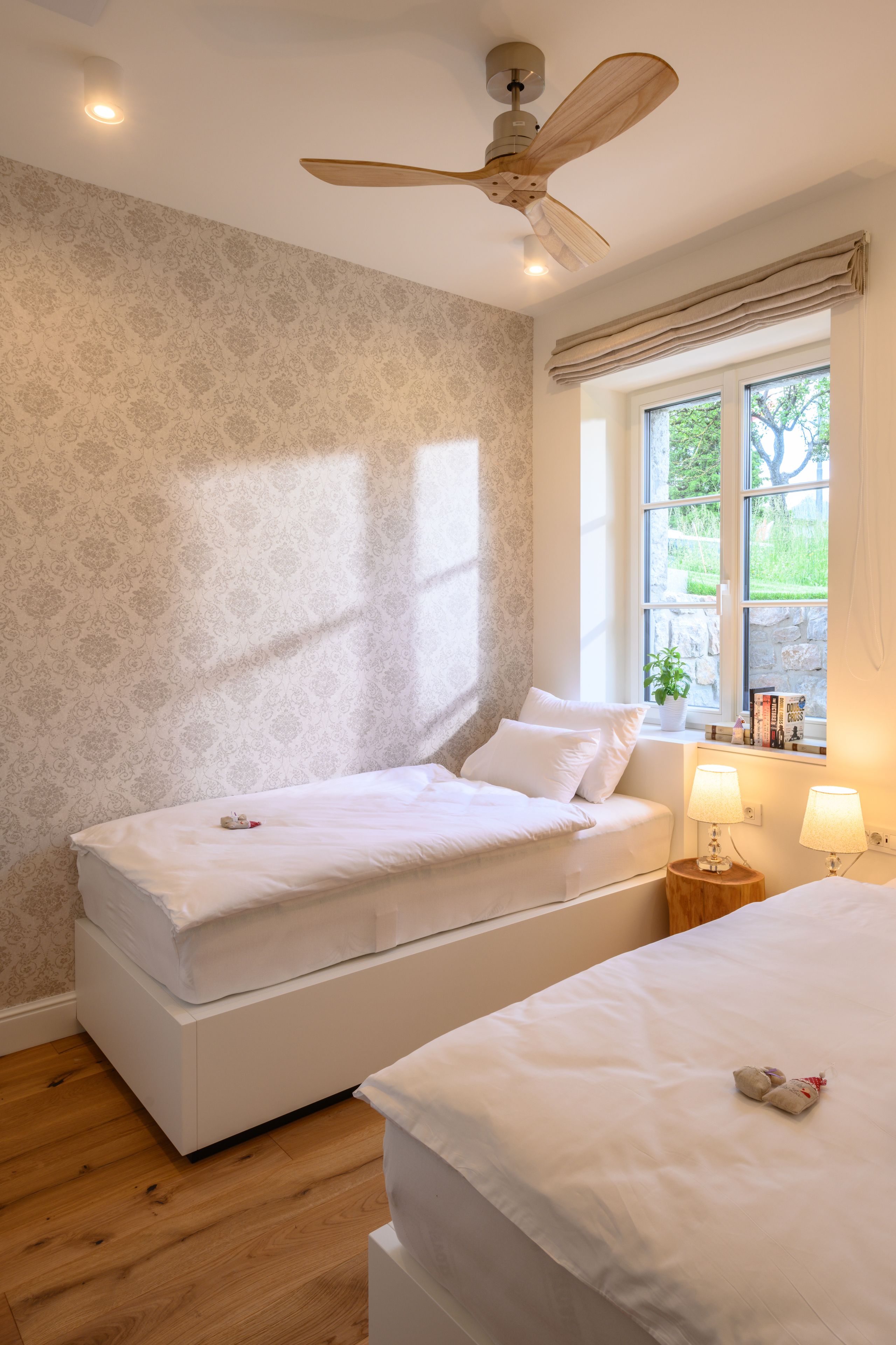 A bedroom with two beds, a ceiling fan, a window with a view of greenery, and a bedside table with lamps.