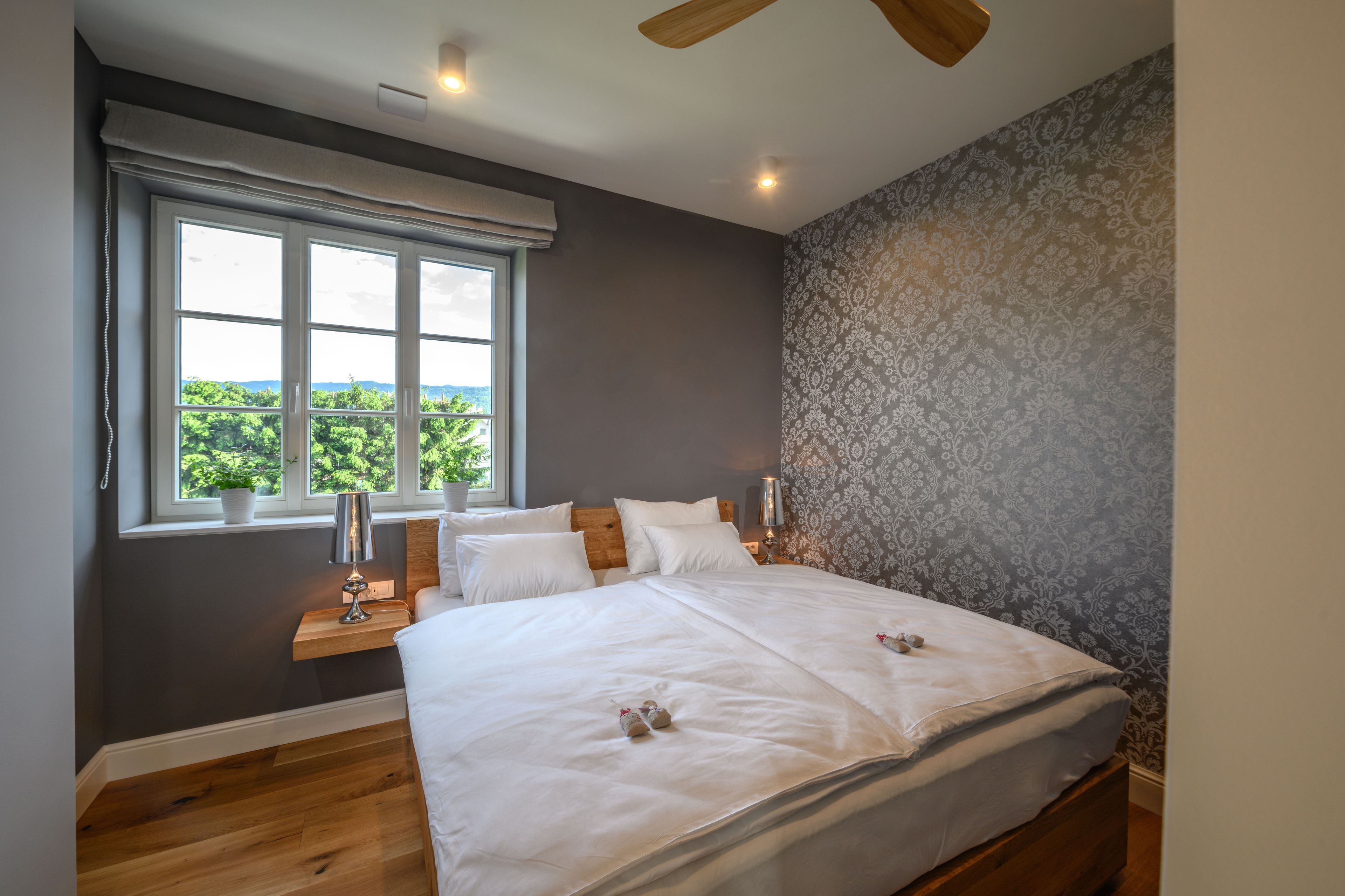 A bedroom with a large bed, a window with a view, a bedside table with a lamp, and a patterned wall.