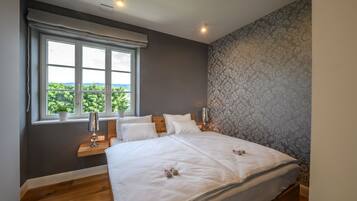 A bedroom with a large bed, a window with a view, a bedside table with a lamp, and a patterned wall.