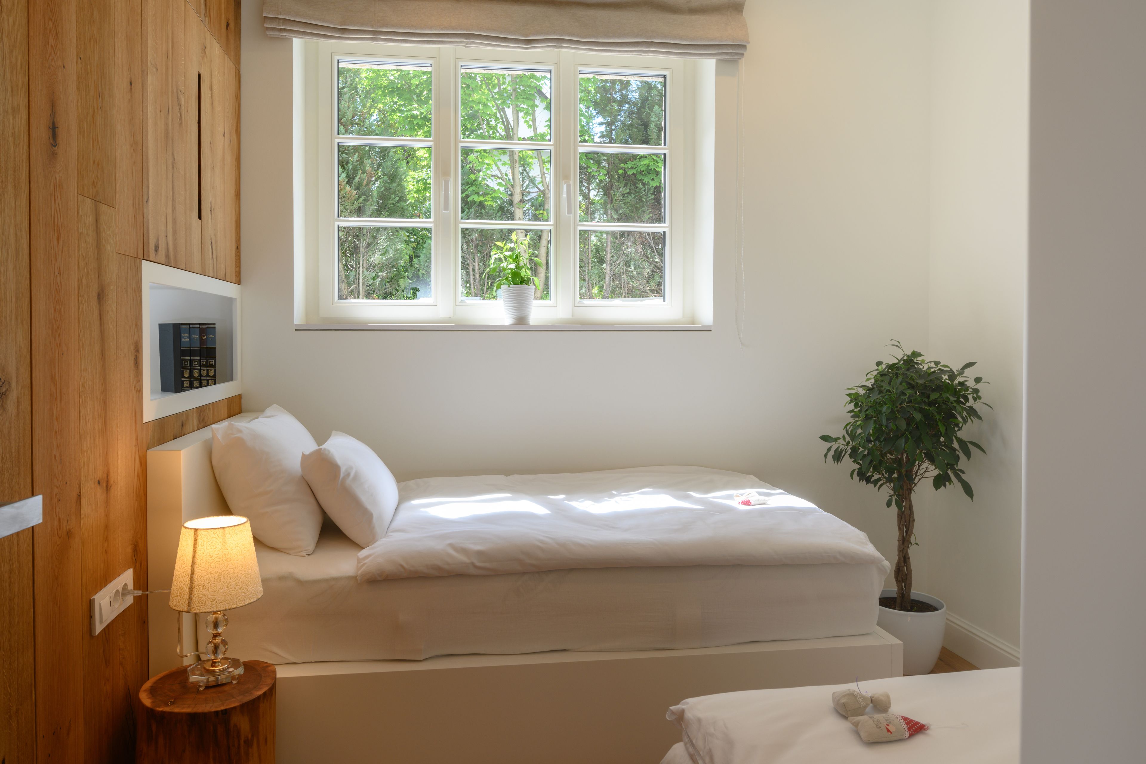 A bedroom with a bed, a bedside lamp, a window with a view of trees, and a potted plant.
