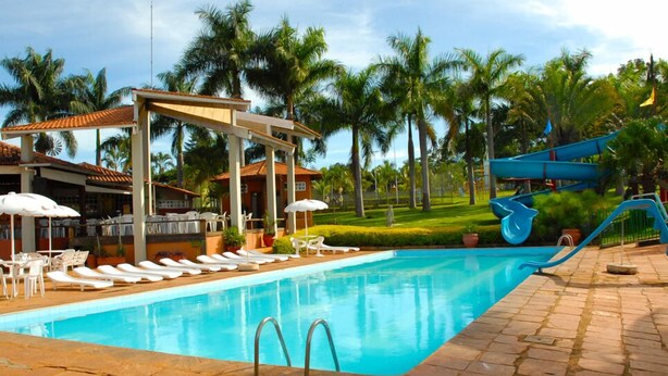 A swimming pool with lounge chairs and umbrellas, a water slide, and palm trees in the background. - Estancia Park Hotel (Anapolis)
