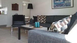 A modern living room with a grey sofa, a black chair, and a coffee table. There is a painting on the wall and a lamp on the floor. - Hals Hotel Apartment (Hals)