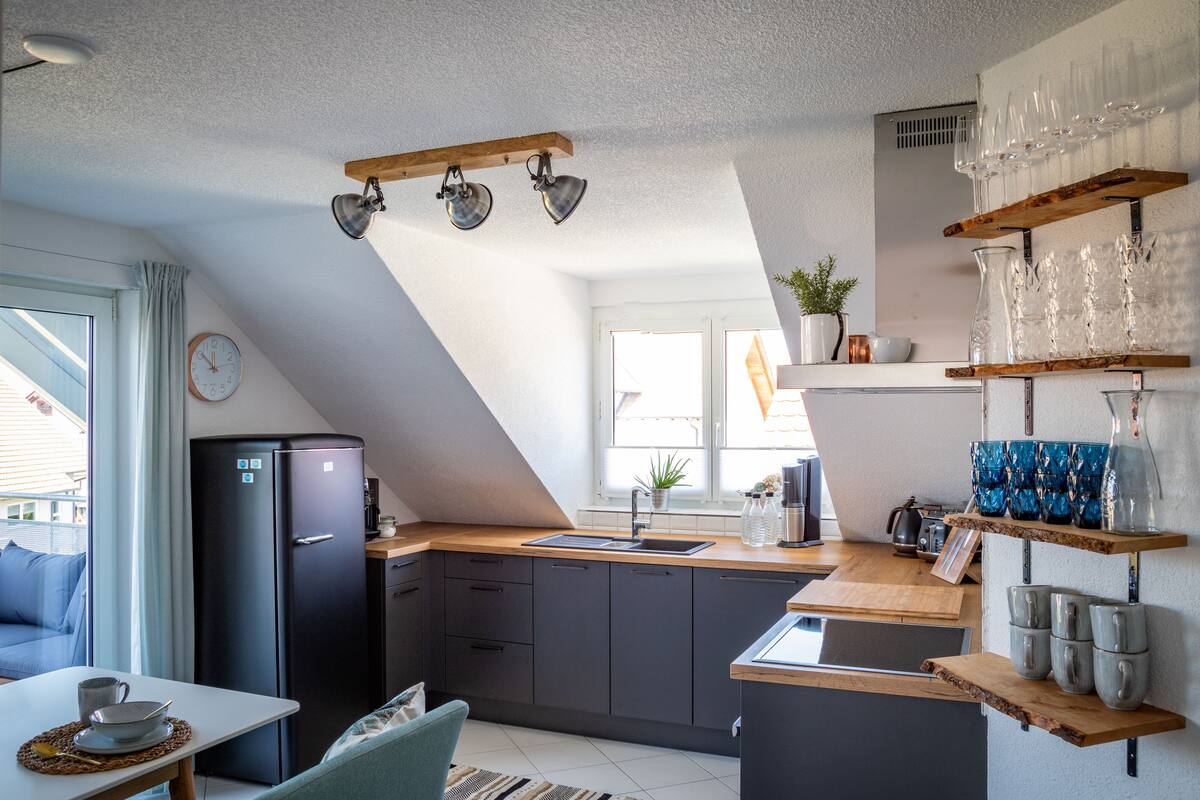 A modern kitchen with a black refrigerator, wooden countertops, and a built-in sink. There are shelves with glassware and a clock on the wall.