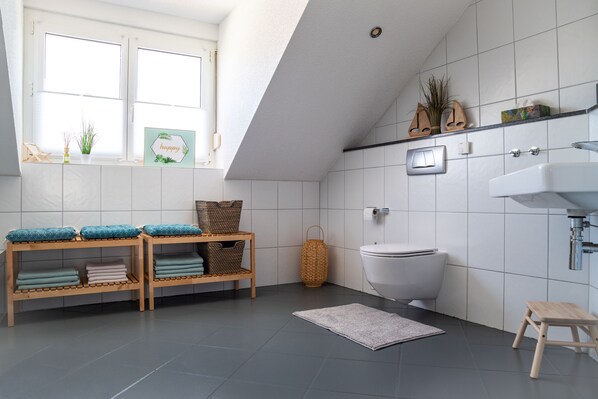A modern bathroom with a white toilet, a sink, and a window. There are shelves with folded towels and a small stool.