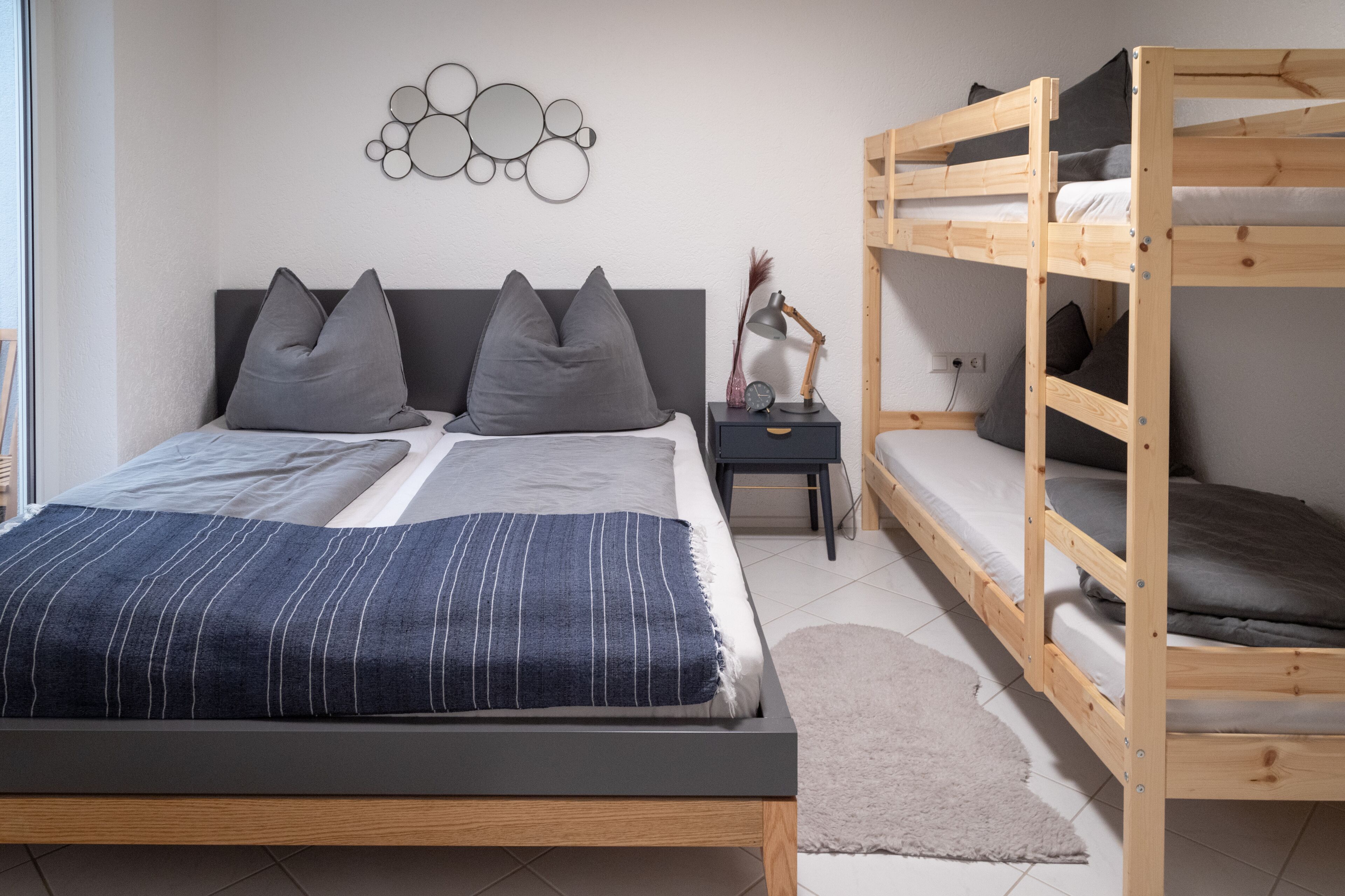 A modern bedroom with a large bed, a smaller bunk bed, a bedside table with a lamp, and a wall art piece.