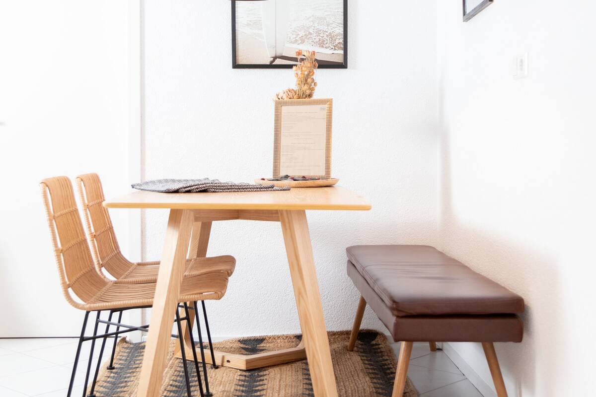 A dining area with a wooden table, two chairs, a bench, a black pendant light, and framed pictures on the wall.