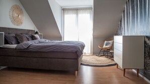 A modern bedroom with a bed, a dresser, a chair, and a large window. - Sohana Lifestyle Apartments (Rust)