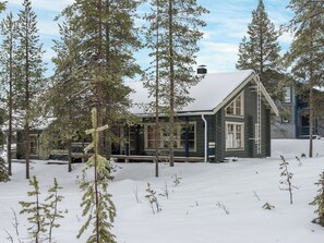 Lumihunnun peittämä mökki, jonka ympärillä kohoaa korkeita, ikivihreitä puita.