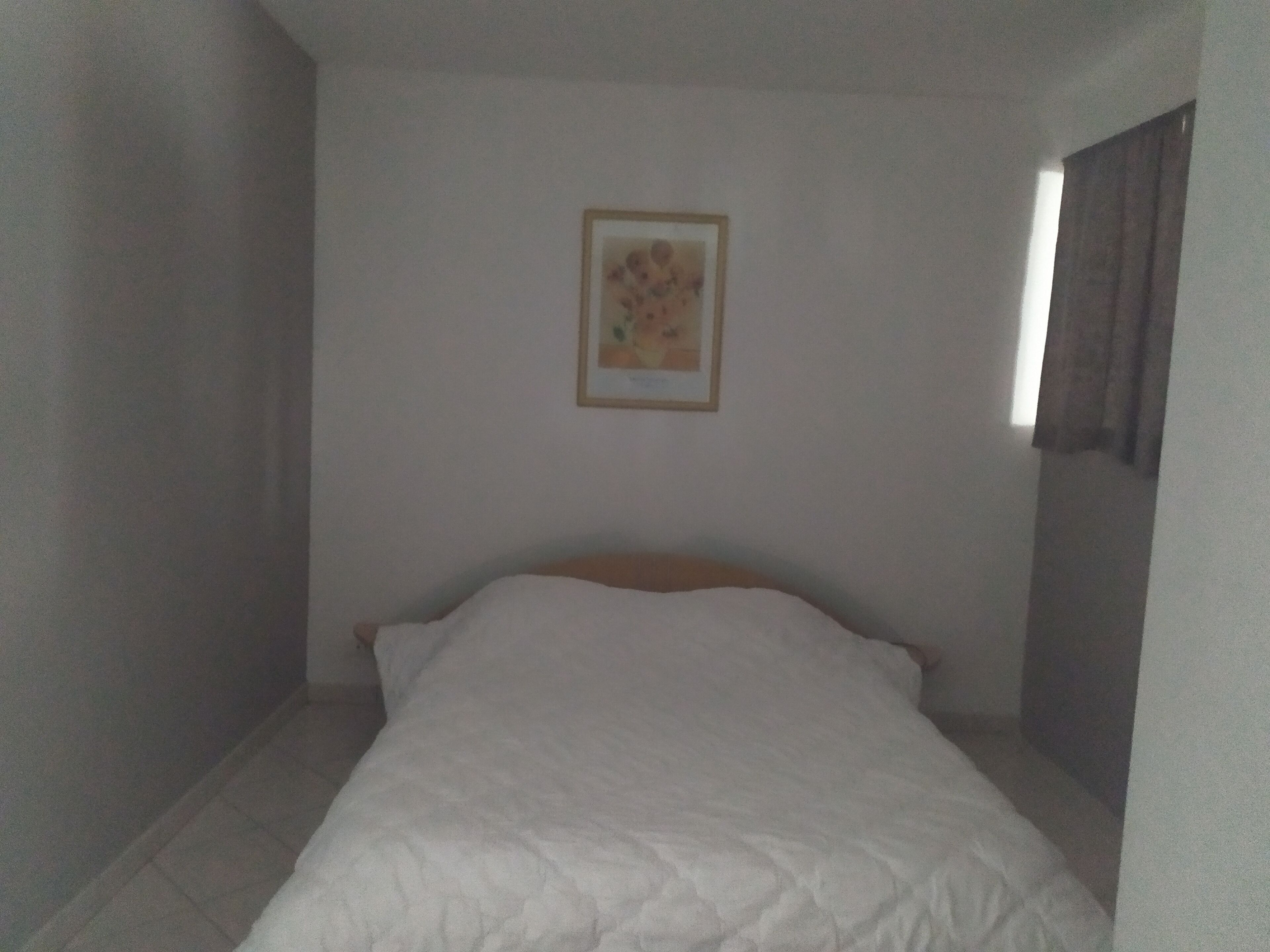 Una stanza piccola e arredata in modo essenziale, con un letto singolo, un quadro appeso e una finestra con tende.
