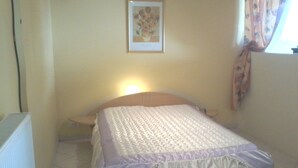 A small, neatly made bed with a white and purple bedspread, a nightstand with a lamp, a framed picture on the wall, and a window with curtains. - rental in the heart of gerardmer (Gérardmer)