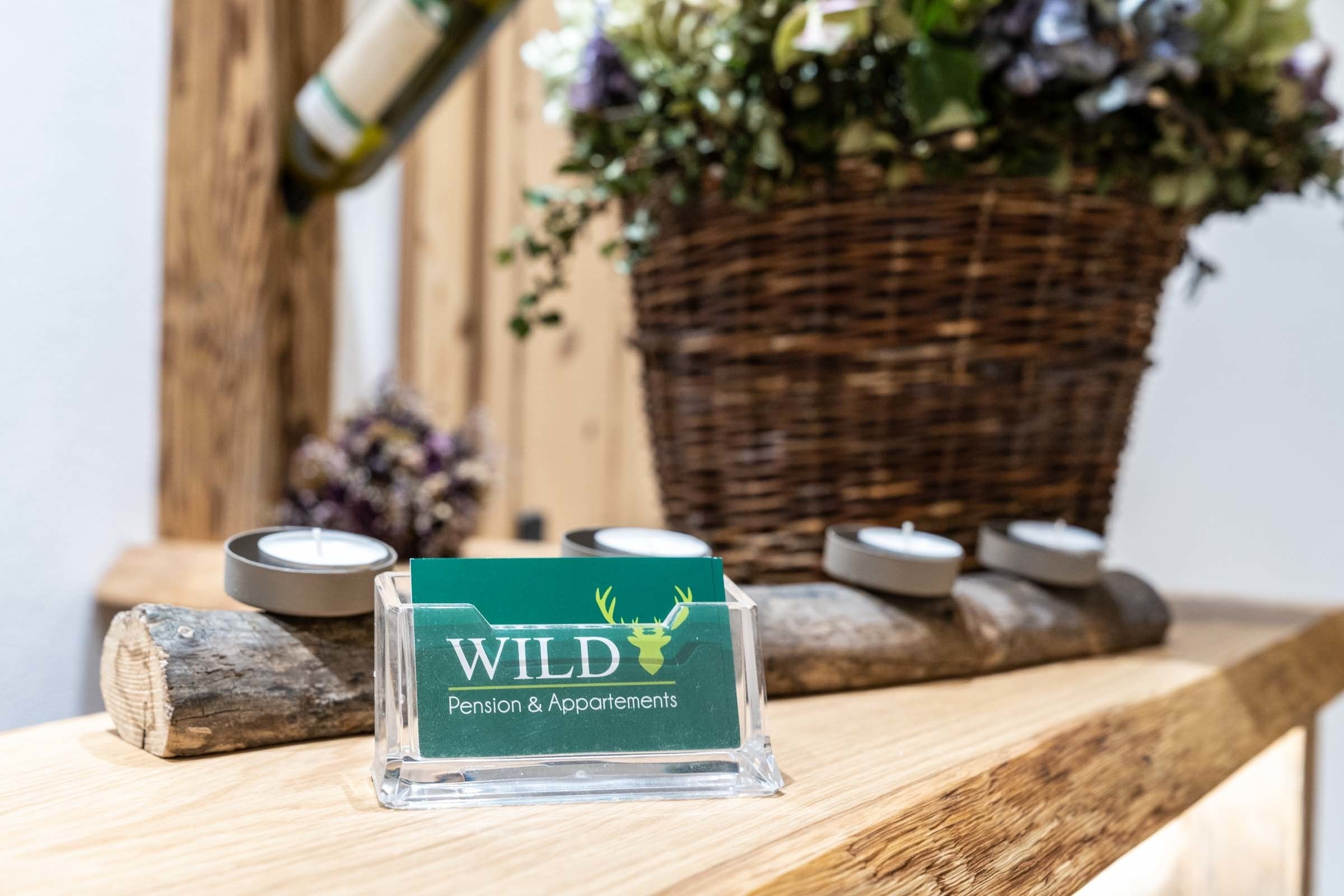 Une plaque transparente en acrylique, portant le logo et le nom « WILD Pension & Apartments », est fixée sur une surface en bois.