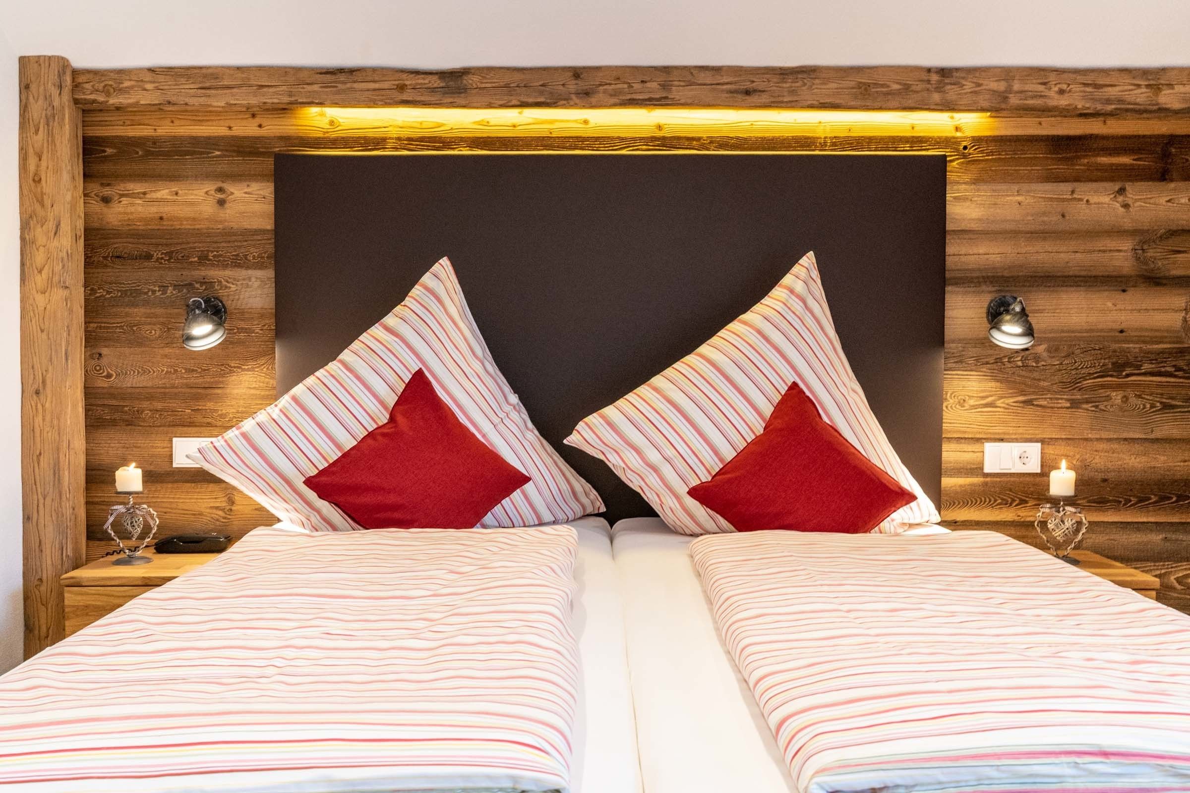A double bed with striped bedding and two red pillows, set against a wooden headboard with a black panel and two wall-mounted lamps.