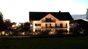 Una casa de dos pisos con un balcón, un porche delantero y un exterior iluminado al atardecer.