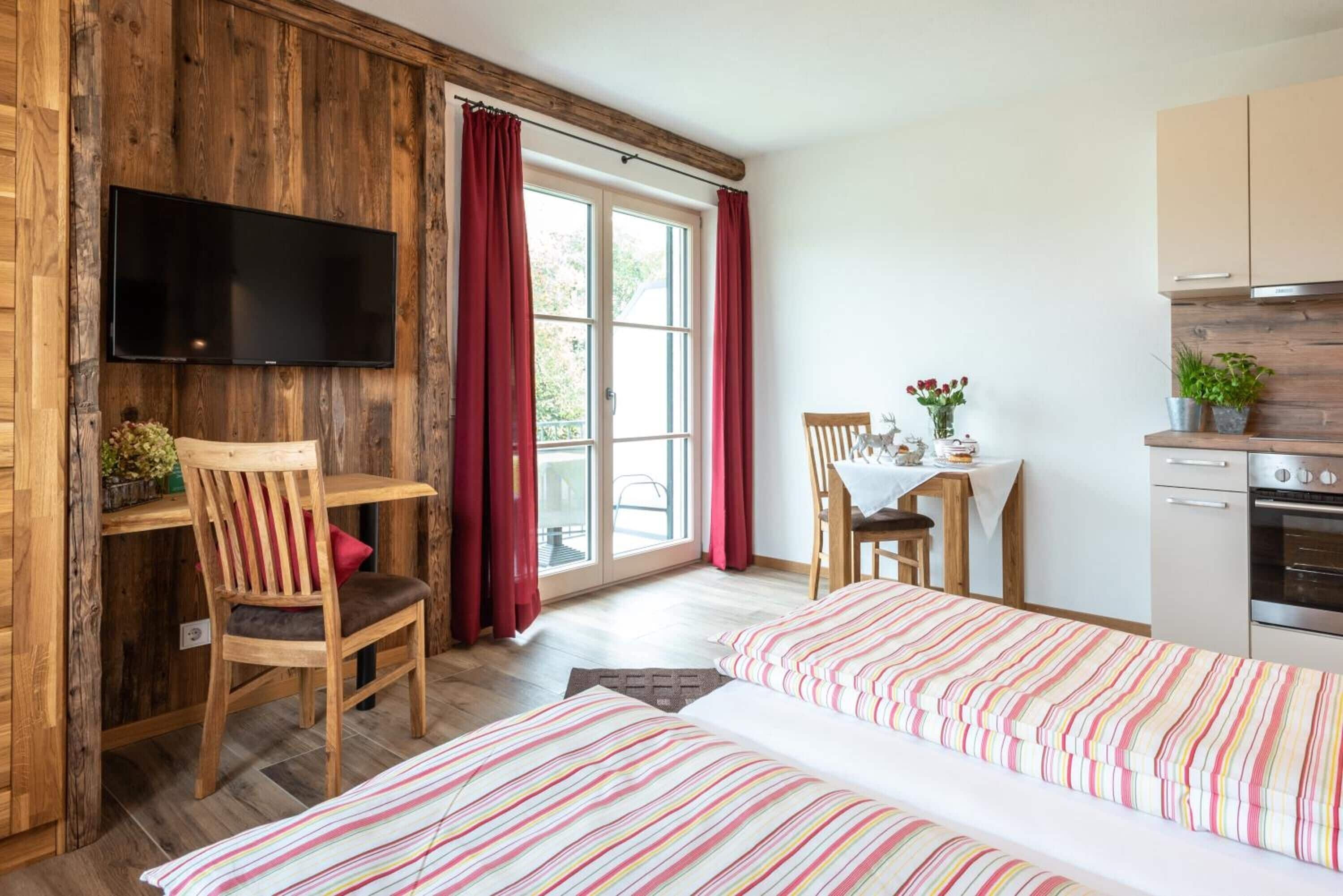 A room with a wooden wall, a TV mounted above a desk, a dining table with chairs, a bed with striped bedding, and a kitchen area with cabinets and an oven.