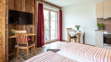 A room with a wooden wall, a TV mounted above a desk, a dining table with chairs, a bed with striped bedding, and a kitchen area with cabinets and an oven.