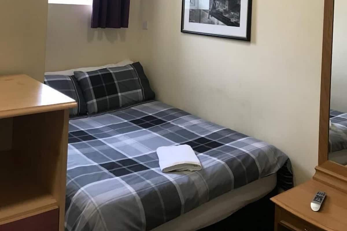 A small, well-lit bedroom with a bed, a bedside table, a mirror, a framed picture on the wall, and a window with curtains.