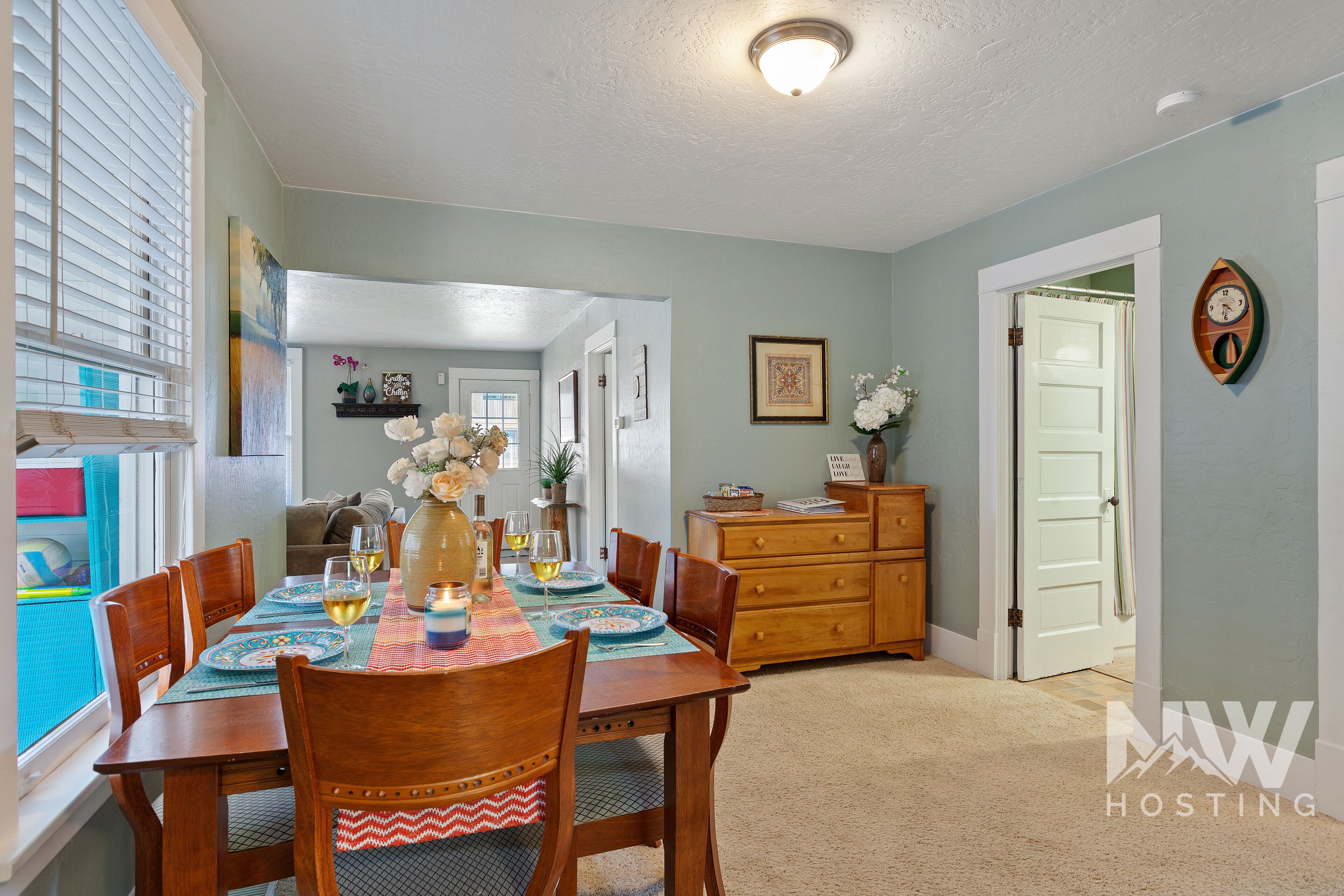 A dining area with a wooden table set for four, a vase with flowers, and a view of a kitchen and living area through an open door.