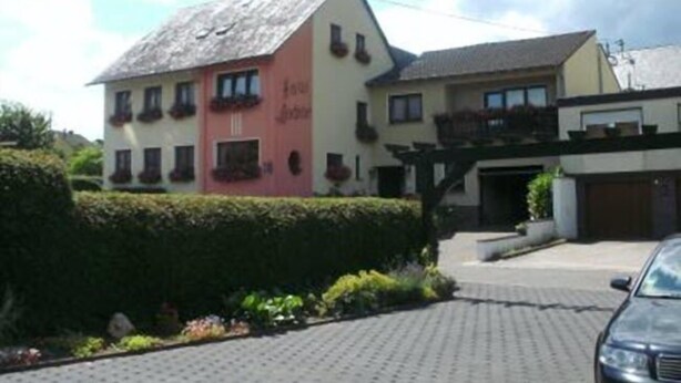 A two-story building with a sloped roof, a balcony on the upper floor, and a paved driveway leading to a parked car. - Gästehaus Andrae (Bruttig-Fankel)