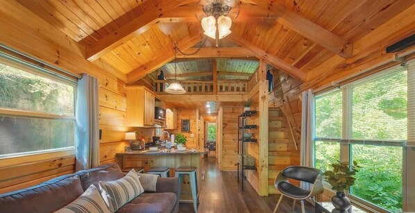 Interior de una cabaña con techo de madera, comedor, cocina y zona de estar con sofá y sillones.