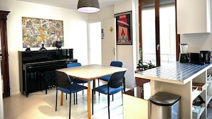 Ett modernt kök med matplats, inrett med ett piano, en stor tavla och en köksö med inbyggda vitvaror.
